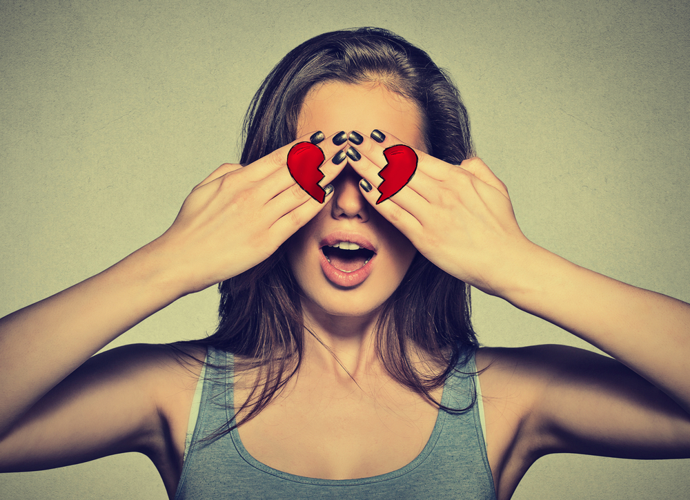 Beautiful young woman eyes covered by hands with broken heart printed on them. Girl in love isolated on gray wall background